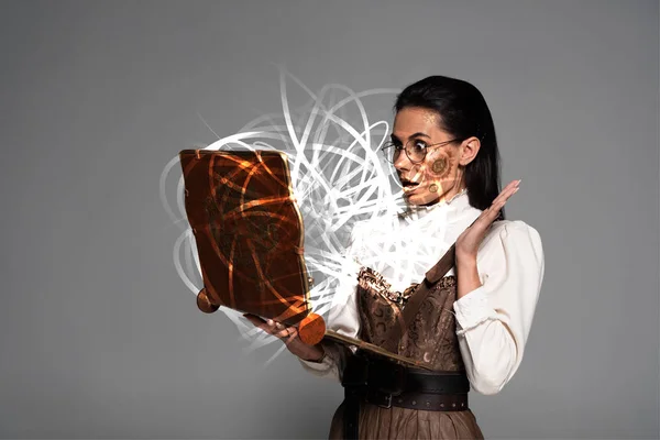 Femme steampunk choqué dans des lunettes en utilisant un ordinateur portable vintage avec une illustration de fée rayonnante isolé sur gris — Photo de stock