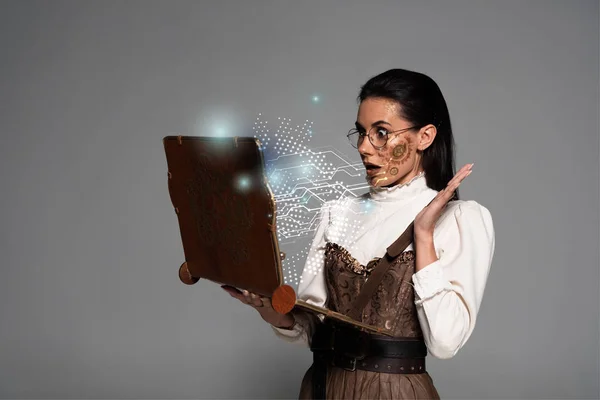 Schockierte Steampunk-Frau in Brille mit Vintage-Laptop mit glühender digitaler Illustration isoliert auf grau — Stockfoto