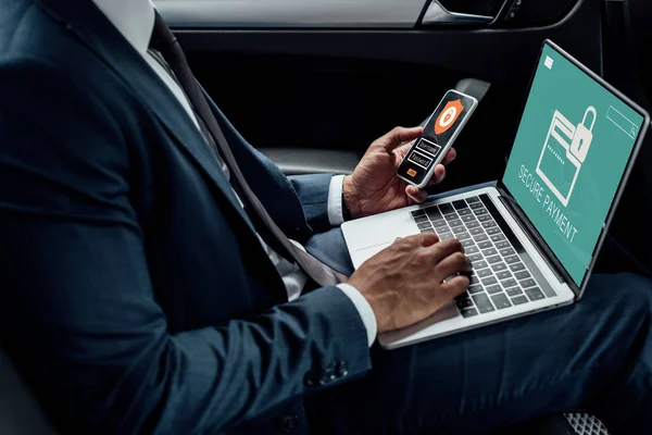 Cropped view of african american businessman using laptop and smartphone in car with cyber security illustration — Stock Photo