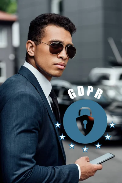 Handsome and confident african american businessman in suit and sunglasses using smartphone with gdpr illustration — Stock Photo