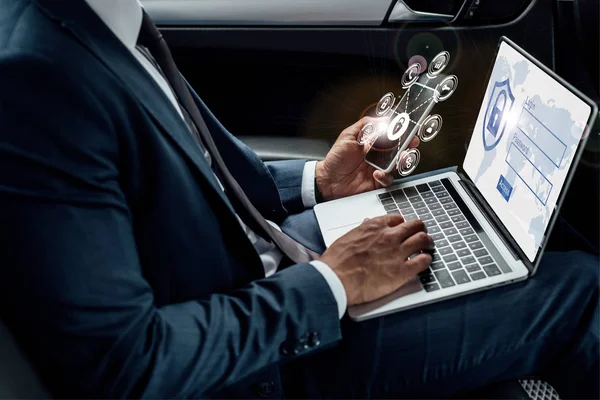 Vista recortada del hombre de negocios afroamericano utilizando el ordenador portátil y el teléfono inteligente en el coche con ilustración de seguridad en Internet - foto de stock