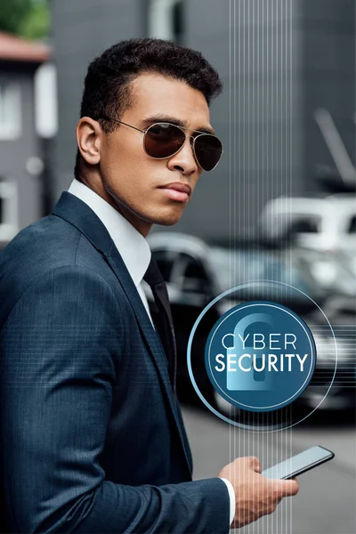 Hombre de negocios afroamericano guapo y confiado en traje y gafas de sol usando teléfono inteligente con ilustración de seguridad cibernética - foto de stock