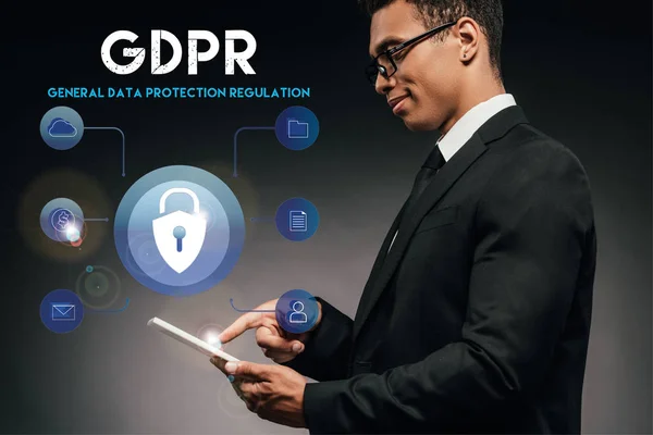 Side view of smiling african american businessman using digital tablet on dark background with gdpr illustration — Stock Photo