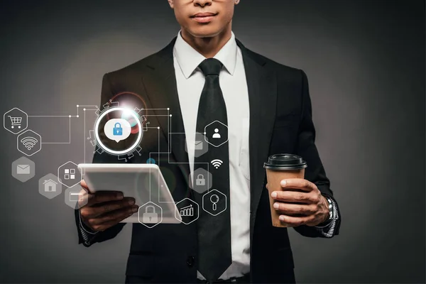 Partial view of african american businessman holding coffee to go and digital tablet on dark background with internet security icons — Stock Photo