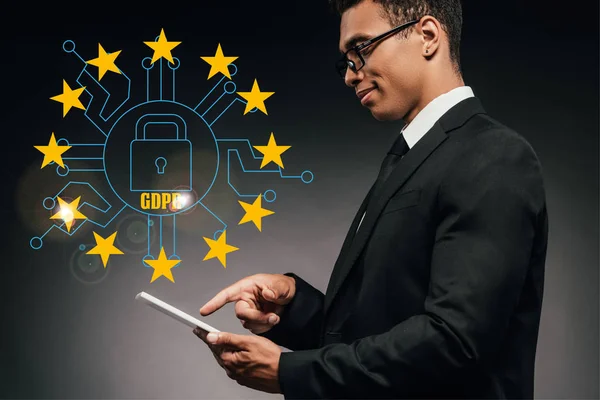 Vista lateral de sonriente hombre de negocios afroamericano utilizando tableta digital sobre fondo oscuro con ilustración gdpr - foto de stock