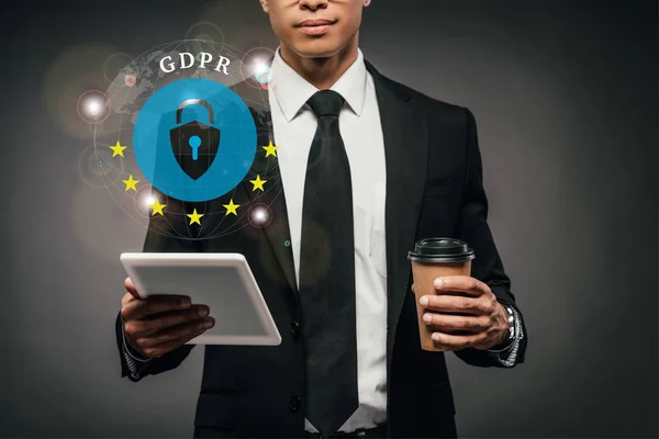 Vista parcial del empresario afroamericano sosteniendo café para llevar y tableta digital sobre fondo oscuro con ilustración gdpr - foto de stock