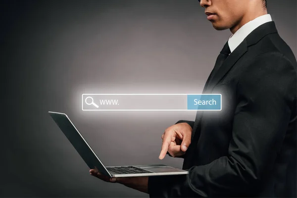 Vista parcial del hombre de negocios afroamericano usando portátil sobre fondo oscuro con ilustración de barra de búsqueda - foto de stock