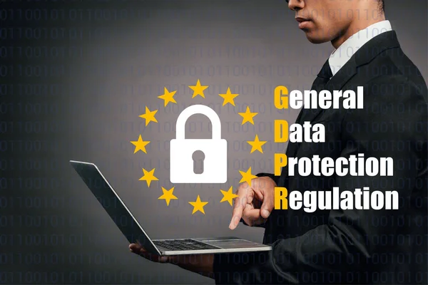 Vista parcial del hombre de negocios afroamericano usando portátil sobre fondo oscuro con ilustración gdpr - foto de stock