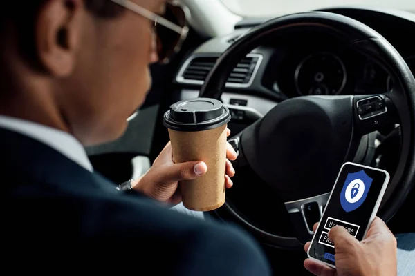 Vista trasera del hombre de negocios afroamericano utilizando un teléfono inteligente con ilustración de acceso seguro y beber café en el coche - foto de stock