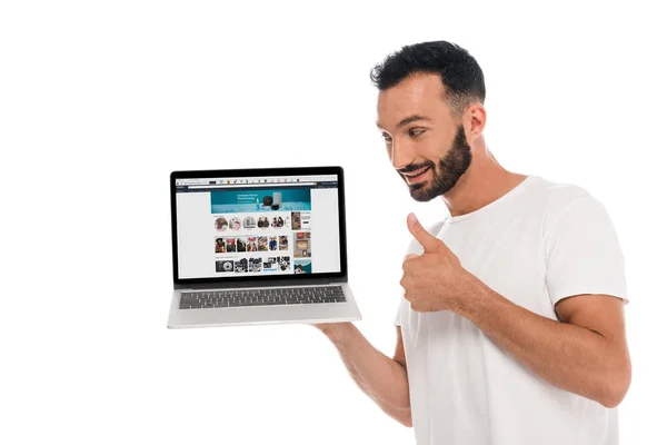 КИЕВ, УКРАИНА - 3 СЕНТЯБРЯ 2019: счастливый бородатый мужчина показывает большой палец вверх и смотрит на ноутбук с Amazon сайт на экране изолированы на белом — стоковое фото