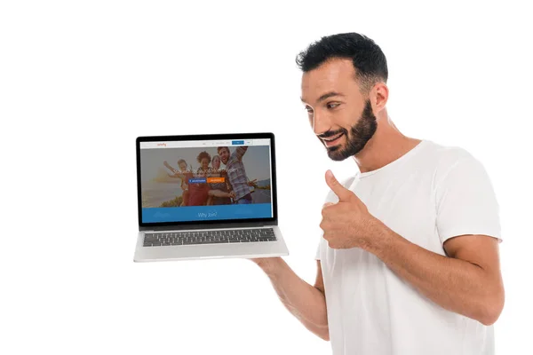 KYIV, UKRAINE - SEPTEMBER 3, 2019: happy bearded man showing thumb up and looking at laptop with couchsurfing website on screen isolated on white — Stock Photo