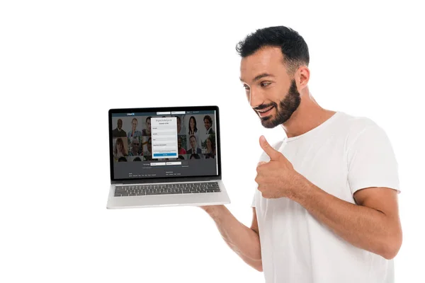 KYIV, UCRANIA - 3 DE SEPTIEMBRE DE 2019: hombre barbudo feliz mostrando el pulgar hacia arriba y mirando a la computadora portátil con linkedin sitio web en la pantalla aislada en blanco - foto de stock