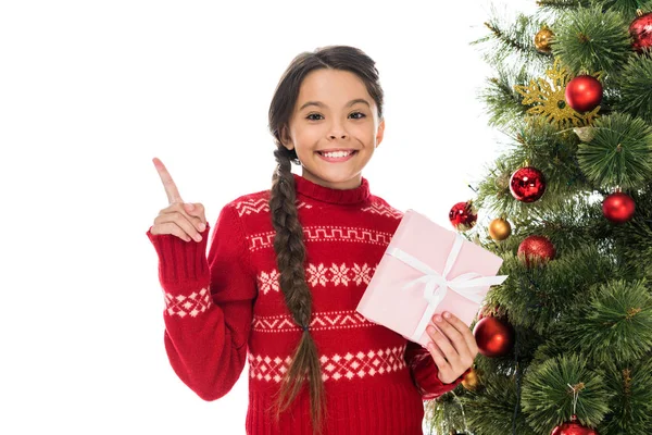 Happy kid holding pink present and pointing with finger near christmas tree isolated on white — Stock Photo