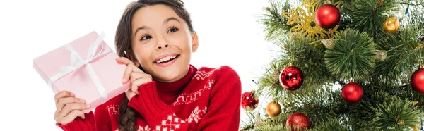 Panoramic shot of smiling kid holding pink present near christmas tree isolated on white — Stock Photo