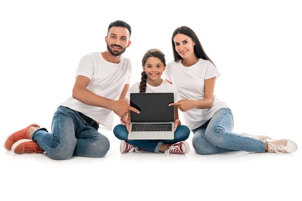 Padres alegres en camisetas blancas que señalan con los dedos a la computadora portátil con pantalla en blanco cerca de hija aislada en blanco - foto de stock