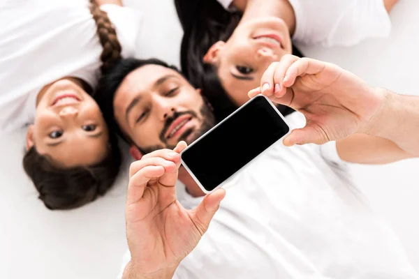 Vista superior del hombre barbudo feliz sosteniendo teléfono inteligente con pantalla en blanco cerca de la esposa y el niño lindo aislado en blanco - foto de stock