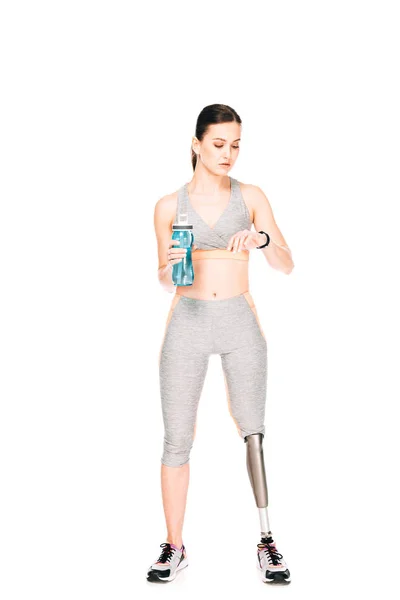 Full length view of sportswoman with prosthetic leg holding sport bottle and looking at smartwatch isolated on white — Stock Photo