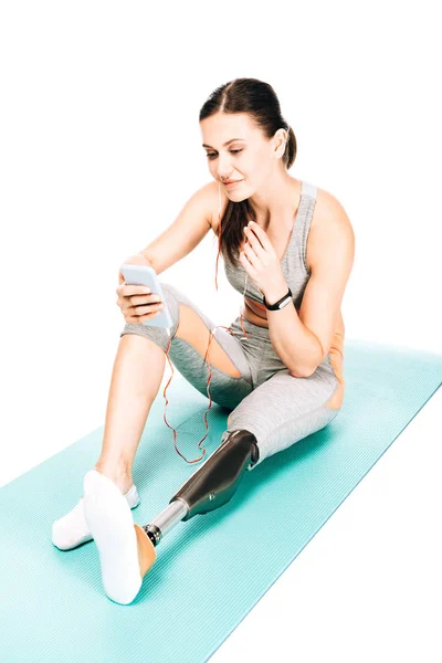 Disabled sportswoman sitting on fitness mat and listening music in earphones isolated on white — Stock Photo