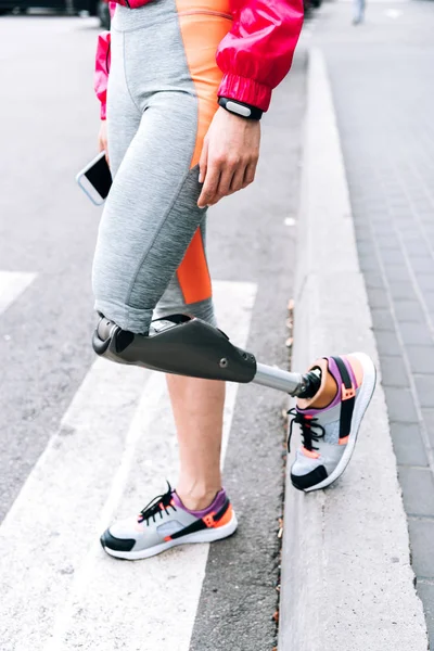 Partial view of disabled sportswoman with prosthesis holding smartphone on street — Stock Photo