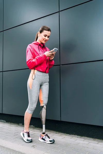 Ganzkörperansicht einer lächelnden behinderten Sportlerin mit Prothese mittels Smartphone auf der Straße — Stockfoto