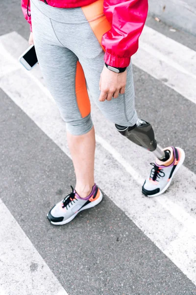 Partial view of disabled sportswoman with prosthesis holding smartphone on street — Stock Photo