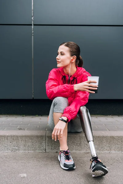 Deportista discapacitada escuchando música en auriculares y sosteniendo la taza en la calle - foto de stock