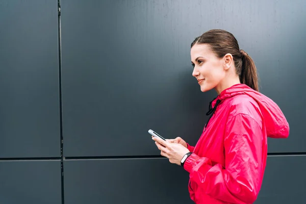 Vue latérale d'une sportive souriante tenant un smartphone dans la rue — Photo de stock