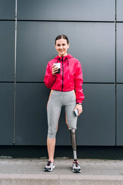 Vista completa de la sonriente deportista discapacitada sosteniendo teléfono inteligente y taza en la calle - foto de stock