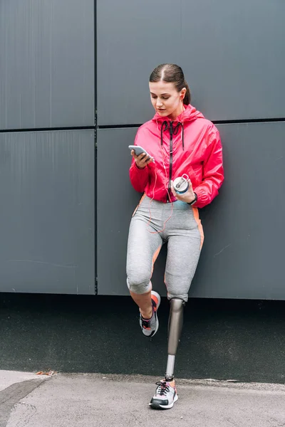 Full length view of disabled sportswoman listening music in earphones and using smartphone on street — Stock Photo