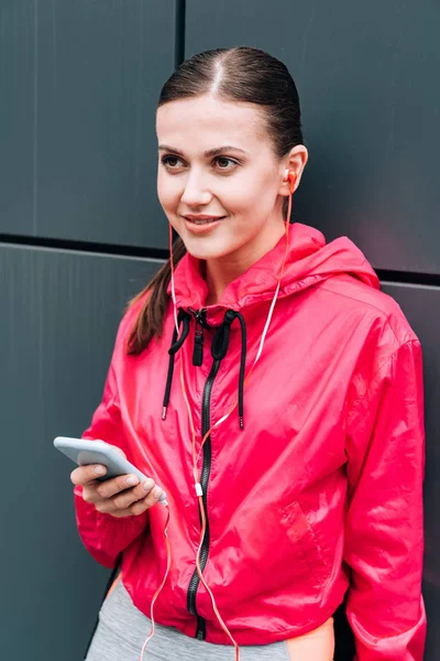 Deportista sonriente escuchando música en auriculares y usando smartphone en la calle - foto de stock
