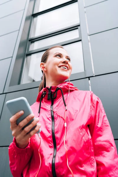 Visão de baixo ângulo de sorrir esportista ouvir música em fones de ouvido e usando smartphone na rua — Fotografia de Stock