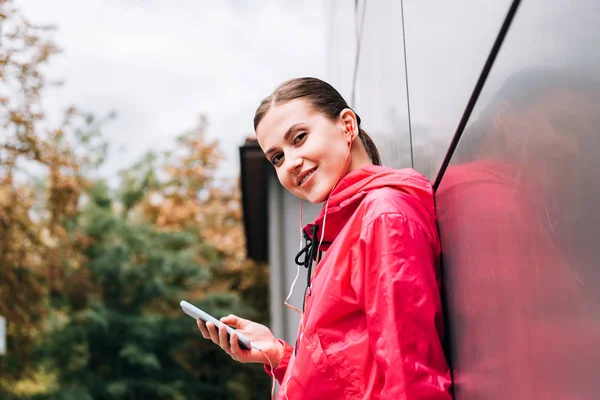 Smiling sportswoman listening music in earphones and using smartphone on street — Stock Photo