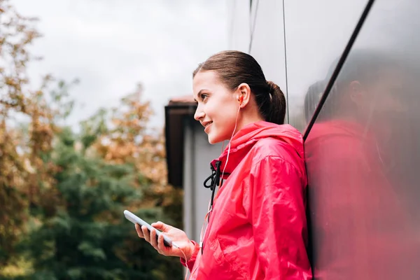 Vue latérale d'une sportive souriante écoutant de la musique dans des écouteurs et utilisant un smartphone dans la rue — Photo de stock