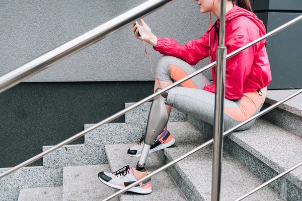 Обрезанный вид спортсменки-инвалида, держащей смартфон и слушающей музыку на лестнице — стоковое фото