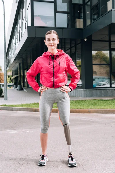 Vista completa de la sonriente deportista discapacitada de pie con las manos en las caderas en la calle - foto de stock