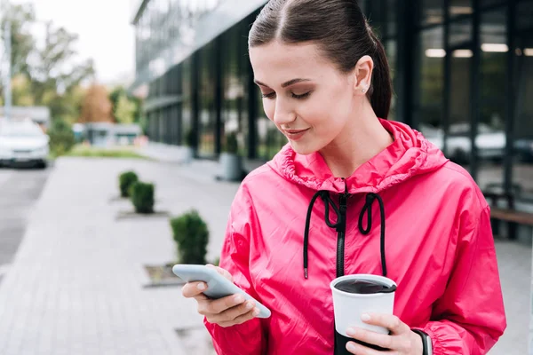 Усміхнена спортсменка тримає чашку і використовує смартфон на вулиці — стокове фото