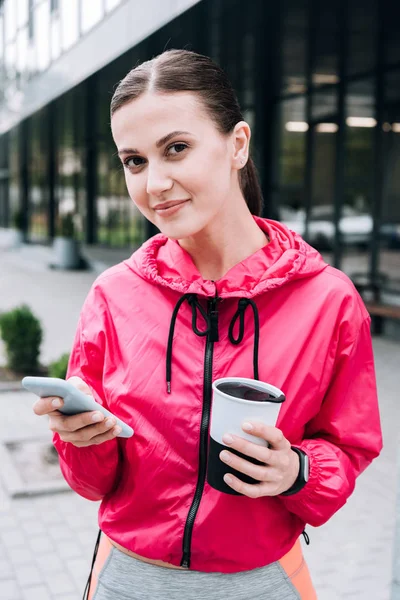 Sportive souriante tenant la tasse et utilisant un smartphone dans la rue — Photo de stock