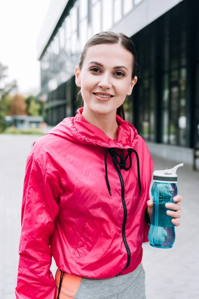 Attrayant sportif souriant tenant bouteille de sport dans la rue — Photo de stock