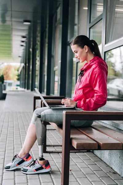 Side view of disabled sportswoman sitting on bench and using laptop on street — Stock Photo