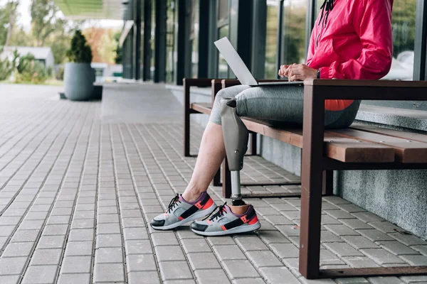 Обрезанный вид спортсменки-инвалида, сидящей на скамейке и использующей ноутбук на улице — стоковое фото