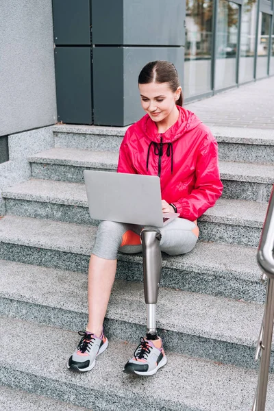 Sorrindo desativado desportista usando laptop enquanto sentado em escadas na rua — Fotografia de Stock