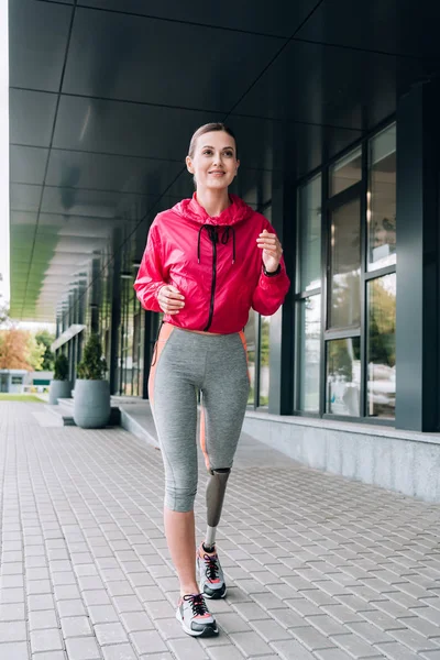 Vista completa de la sonriente deportista discapacitada corriendo por la calle - foto de stock