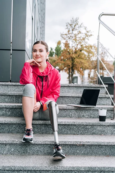 Sorrindo desativado desportista com laptop sentado em escadas na rua — Fotografia de Stock