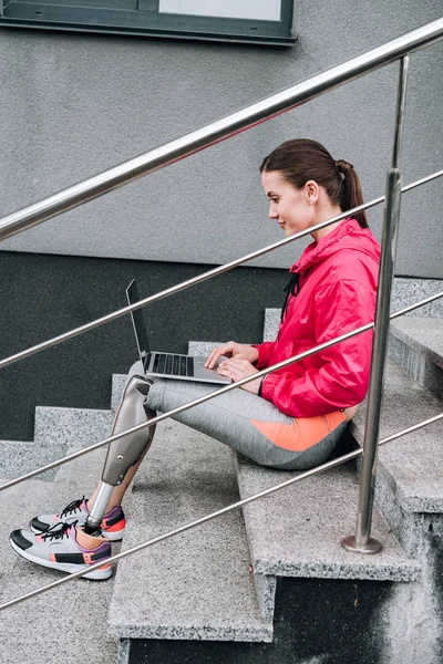 Seitenansicht einer behinderten Sportlerin mit Laptop, während sie auf einer Treppe auf der Straße sitzt — Stockfoto