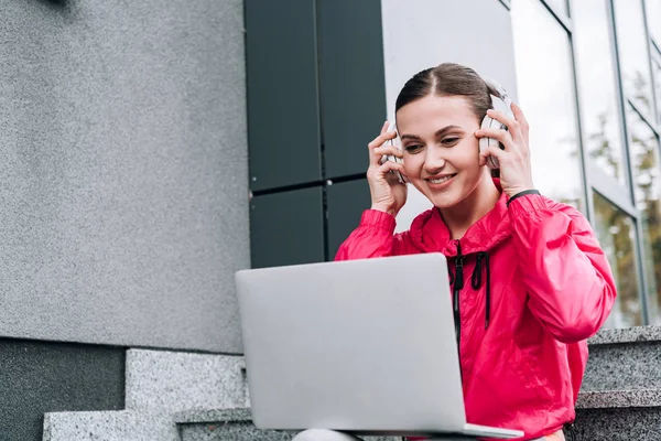 Smiling sportswoman listening music in headphones and using laptop on street — Stock Photo