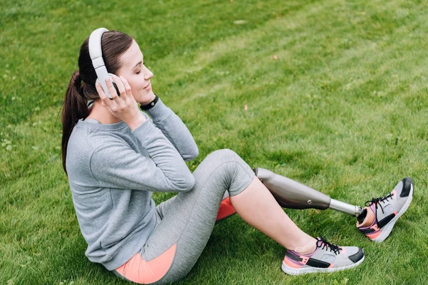 Vista lateral de la deportista discapacitada sentada en la hierba y escuchando música en auriculares - foto de stock