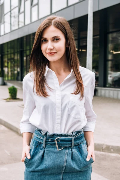 Attractive young woman in white shirt standing with hands in pockets on street — Stock Photo