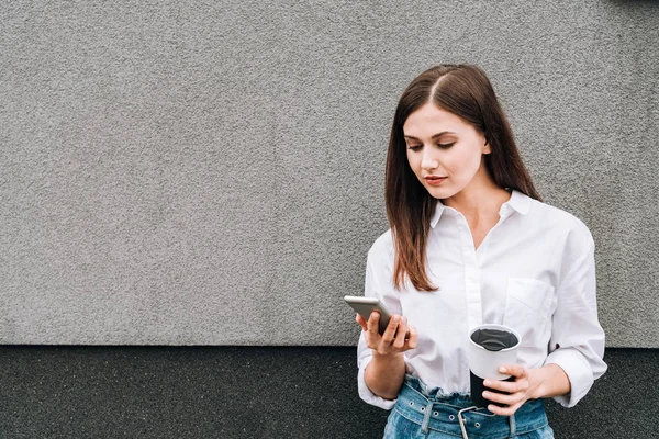 Beautiful young woman holding smartphone and cup on street — Stock Photo