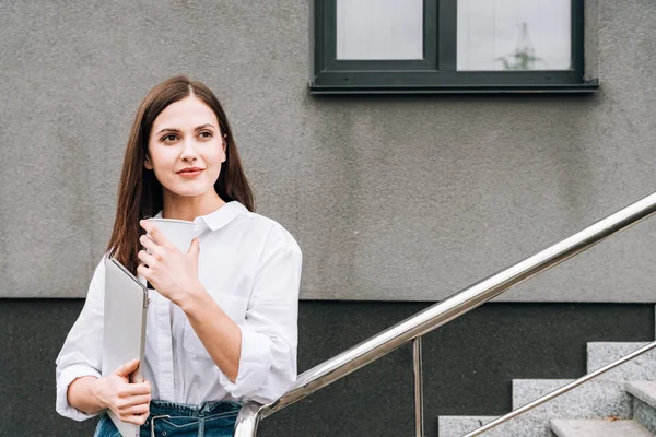 Attraente giovane donna in camicia bianca che tiene il computer portatile sulla strada — Foto stock