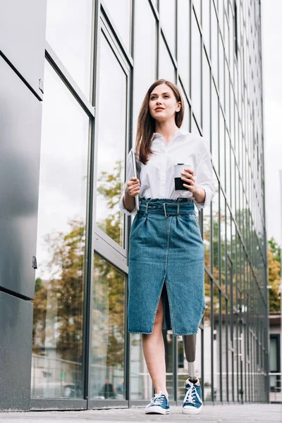 Вид в полный рост женщины-инвалида в джинсовой юбке, держащей ноутбук на улице — стоковое фото
